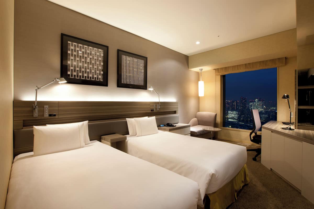Een hotelkamer met twee bedden, een bureau, een stoel en een groot raam met uitzicht op de stad.