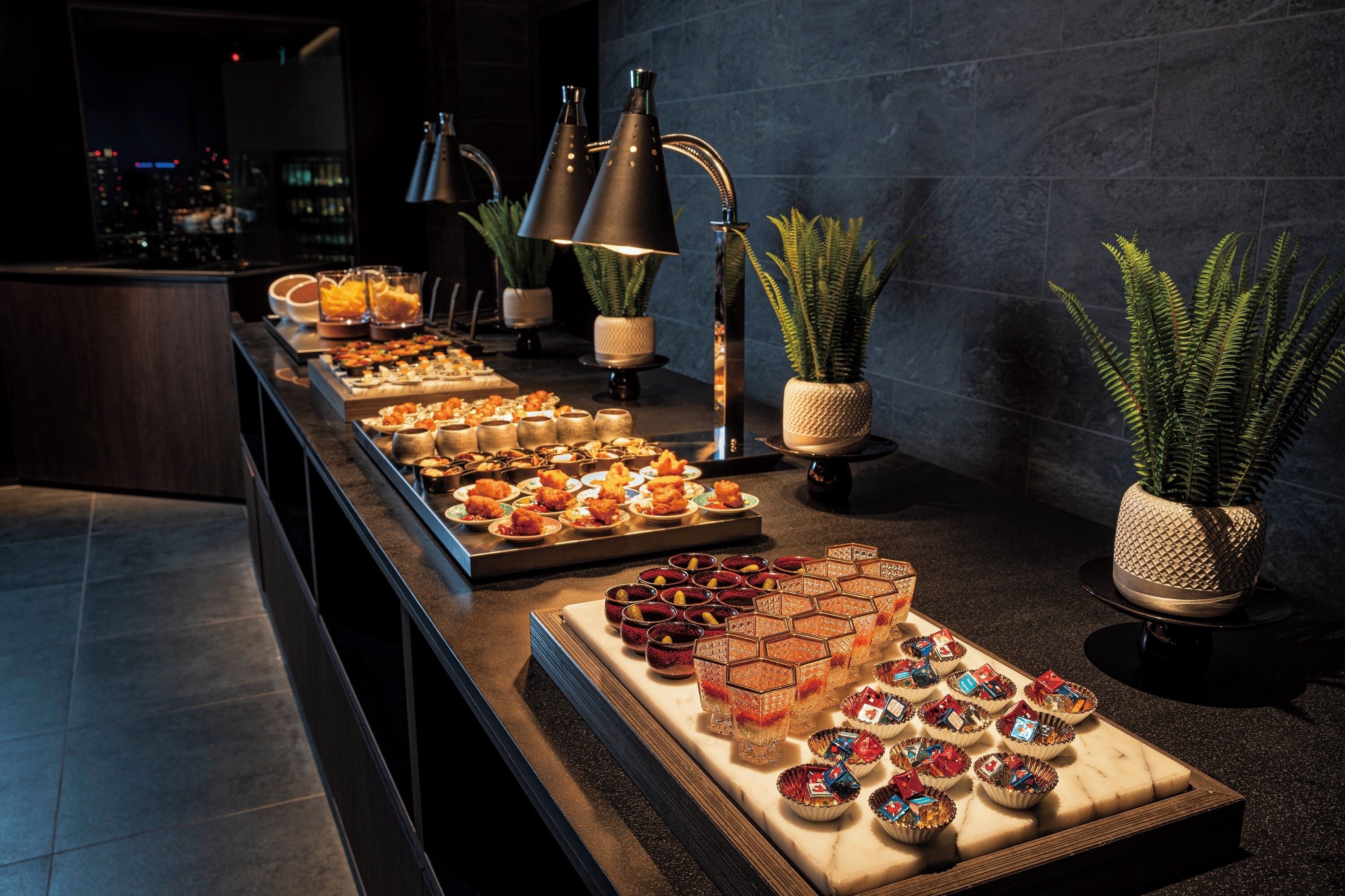 A buffet setup with various food items and drinks, accompanied by potted plants and modern lighting fixtures.