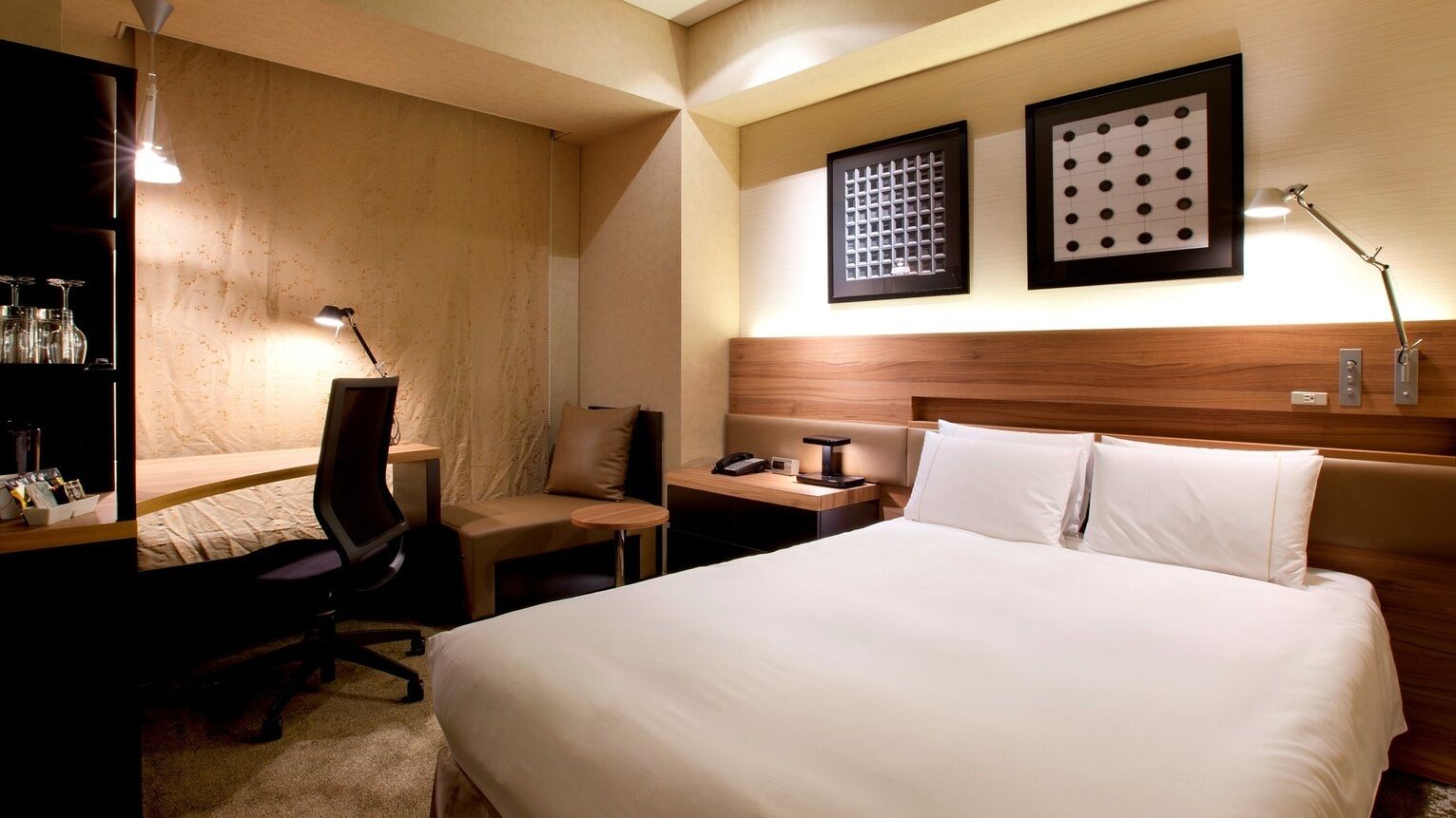 Een hotelkamer met een bed, een bureau met stoel, nachtkastje, lamp en twee ingelijste foto's aan de muur.