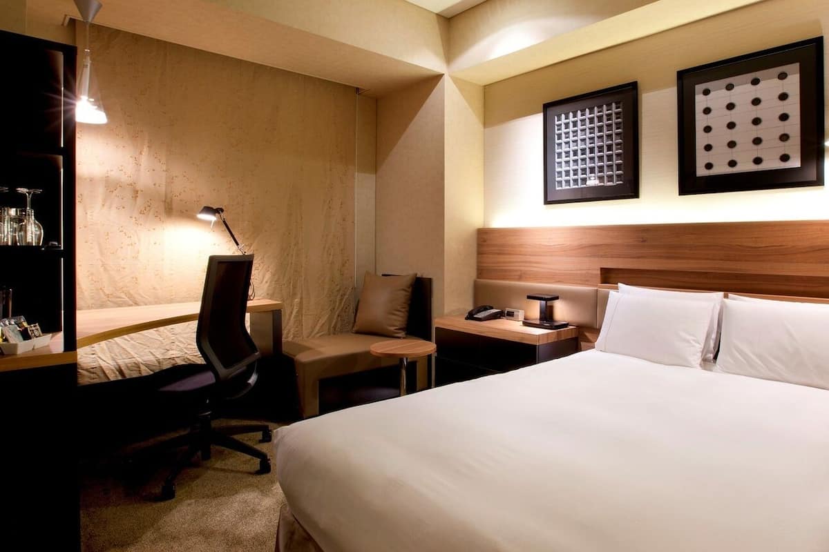 Een hotelkamer met een bed, een bureau met stoel, nachtkastje, lamp en twee ingelijste foto's aan de muur.