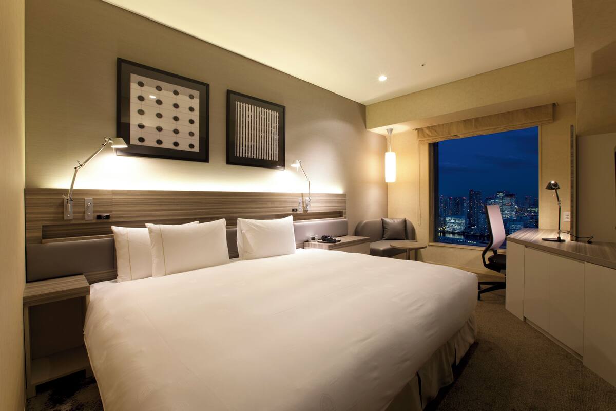 Hotelkamer met een groot bed, nachtkastjes, een bureau, een stoel en uitzicht op een stadsbeeld bij nacht.