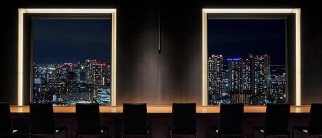 A conference room with a cityscape view, featuring a long table with chairs and a large window.