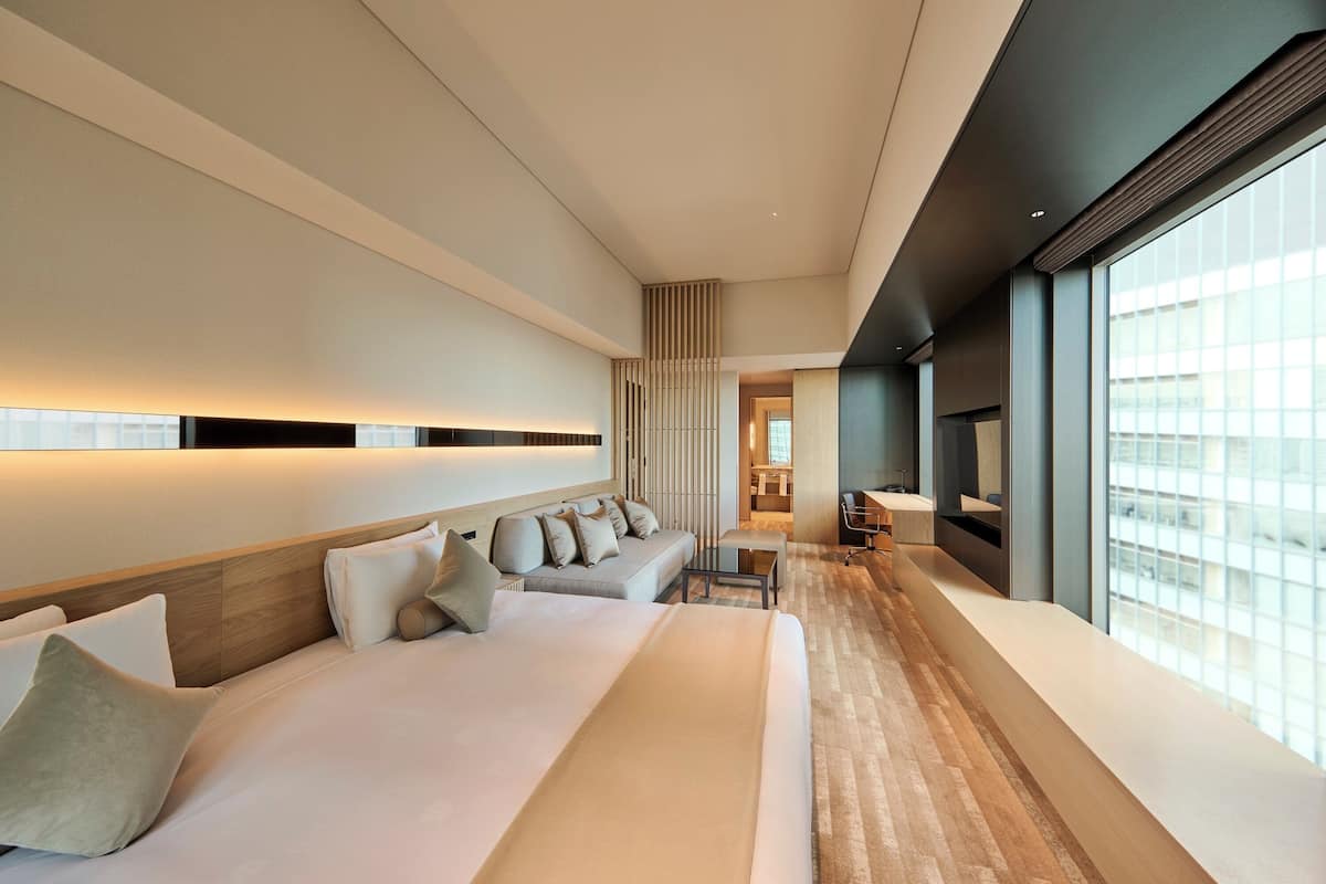 Een moderne hotelkamer met een groot bed, een zithoek en uitzicht op de stad door grote ramen.