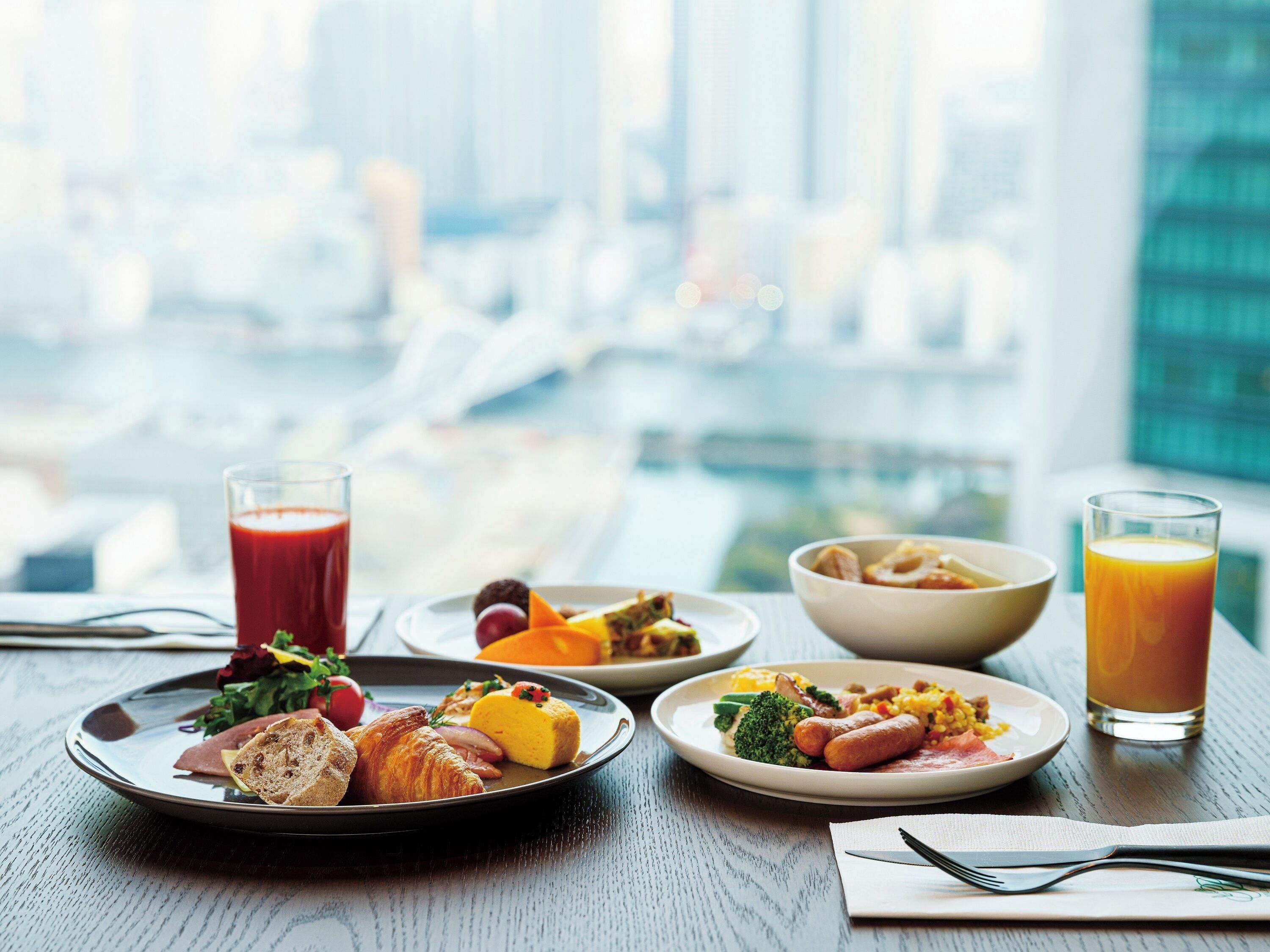 A breakfast setting with croissants, a fruit salad, and a bowl of cereal on a table with a city view in the background.