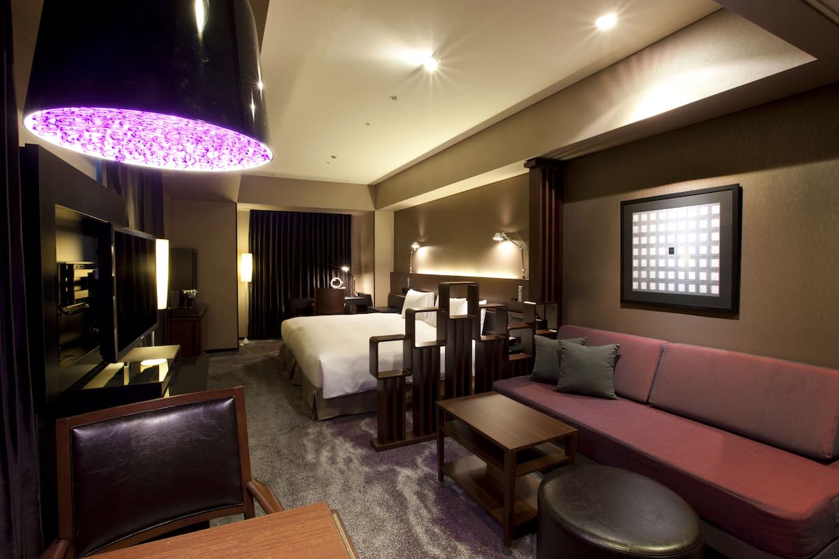 Hotelkamer met een bed, een slaapbank, een bureau, een stoel, een televisie en een grote paarse hanglamp.
