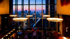 Een modern restaurant met een panoramisch stadszicht door grote ramen, met meerdere hanglampen en diverse zithoeken.