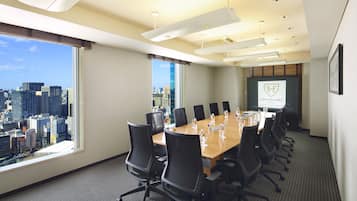 Un salón de conferencias con vistas a la ciudad, una larga mesa con sillas y una pantalla para presentaciones.