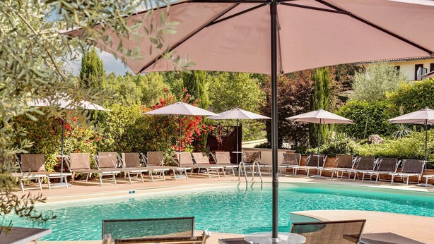 A poolside area with lounge chairs and umbrellas, surrounded by lush greenery and a clear pool. - Hotel El Patio (Corciano)