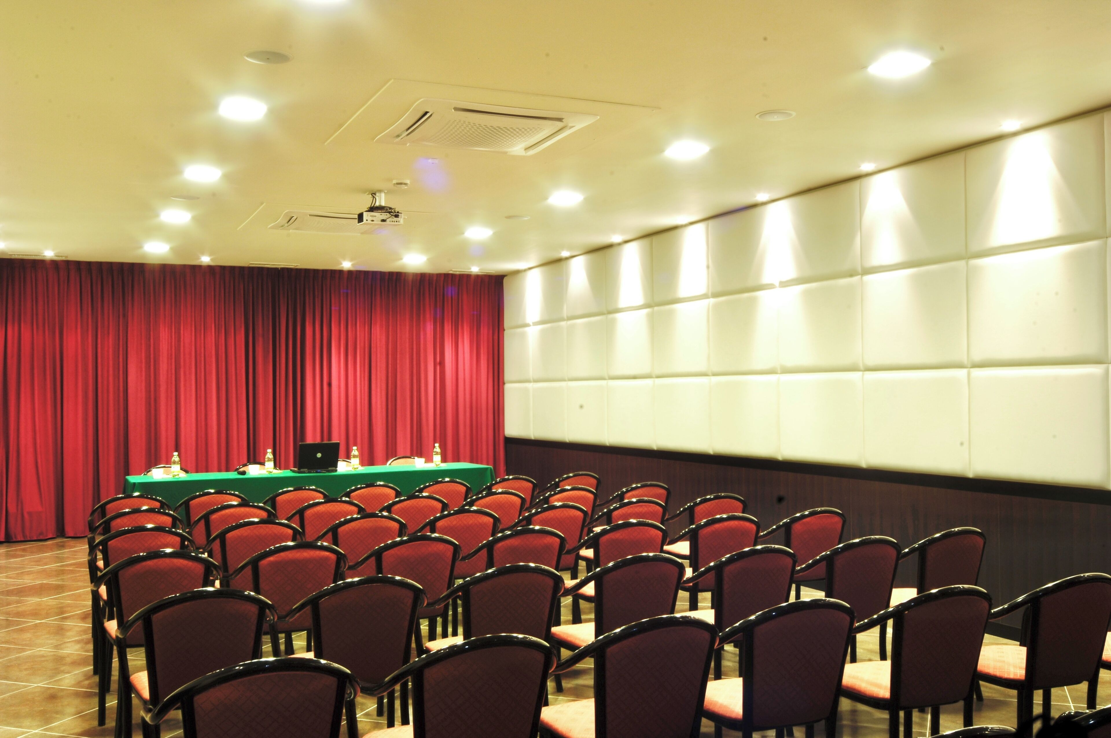 A conference room with rows of red chairs, a green table with a laptop, and a red curtain backdrop.