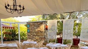 An outdoor patio area with white tables and chairs, a stone wall, and potted plants. - Hotel El Patio (Corciano)