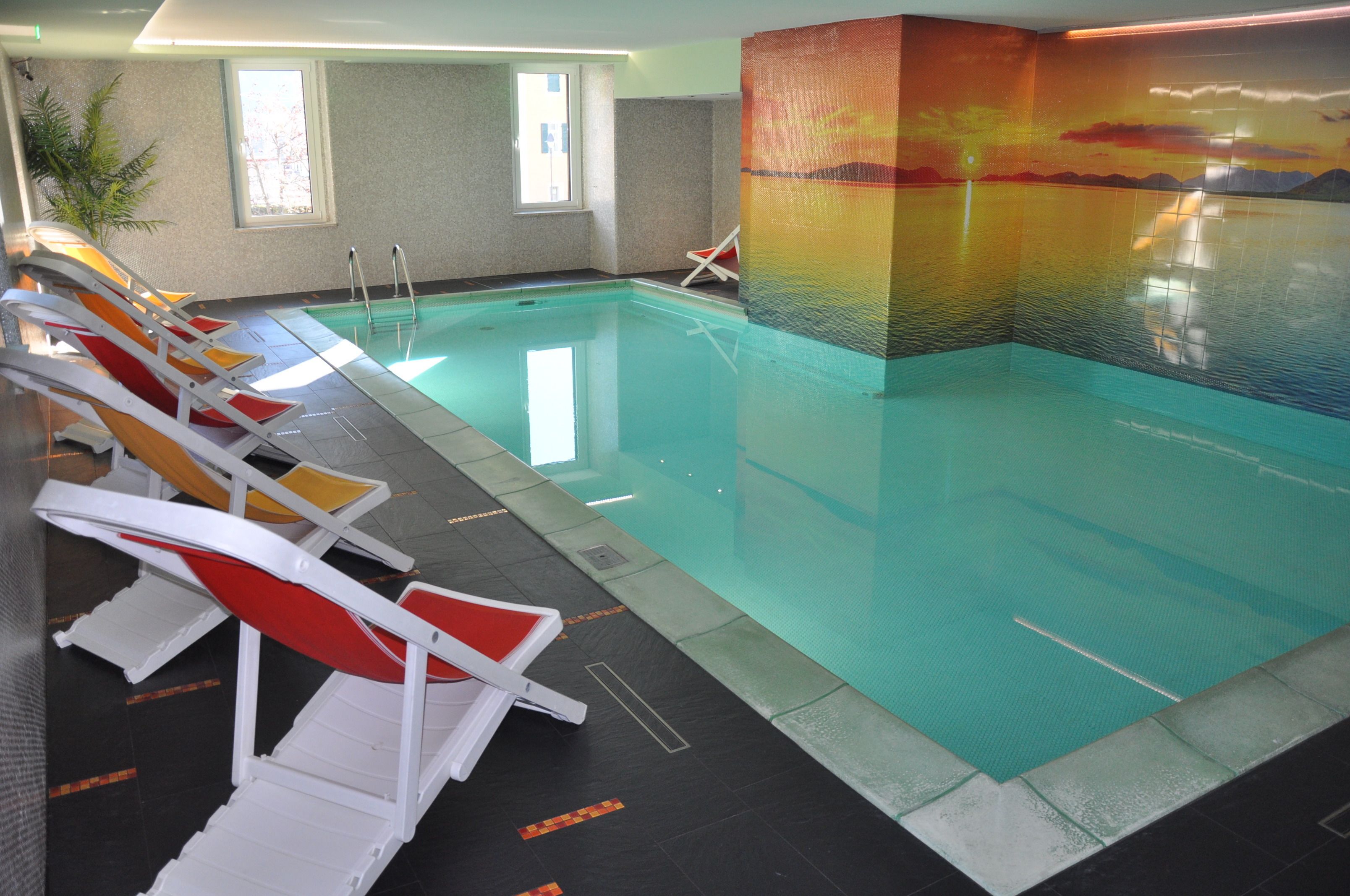 Una piscina interior con varios sillones de sol y un mural que representa un atardecer.