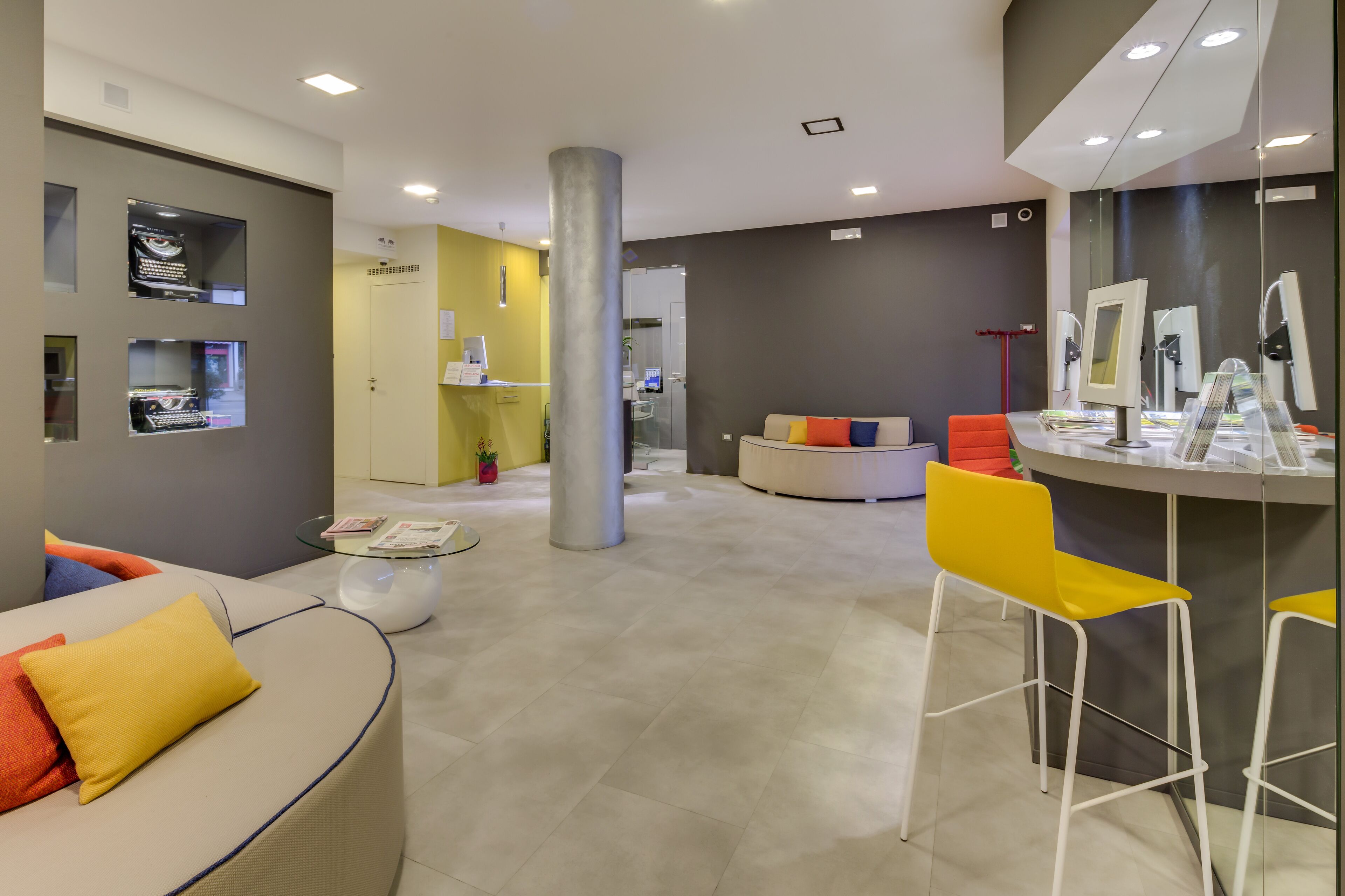 A modern hotel lobby with a curved sofa, a yellow chair, and a reception desk with a computer.
