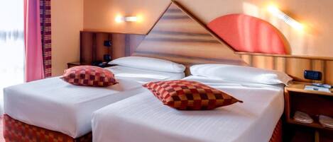 Habitación de hotel con dos camas, cada una con un cojín a cuadros, una mesita de noche con una lámpara y un ventilador de pared.