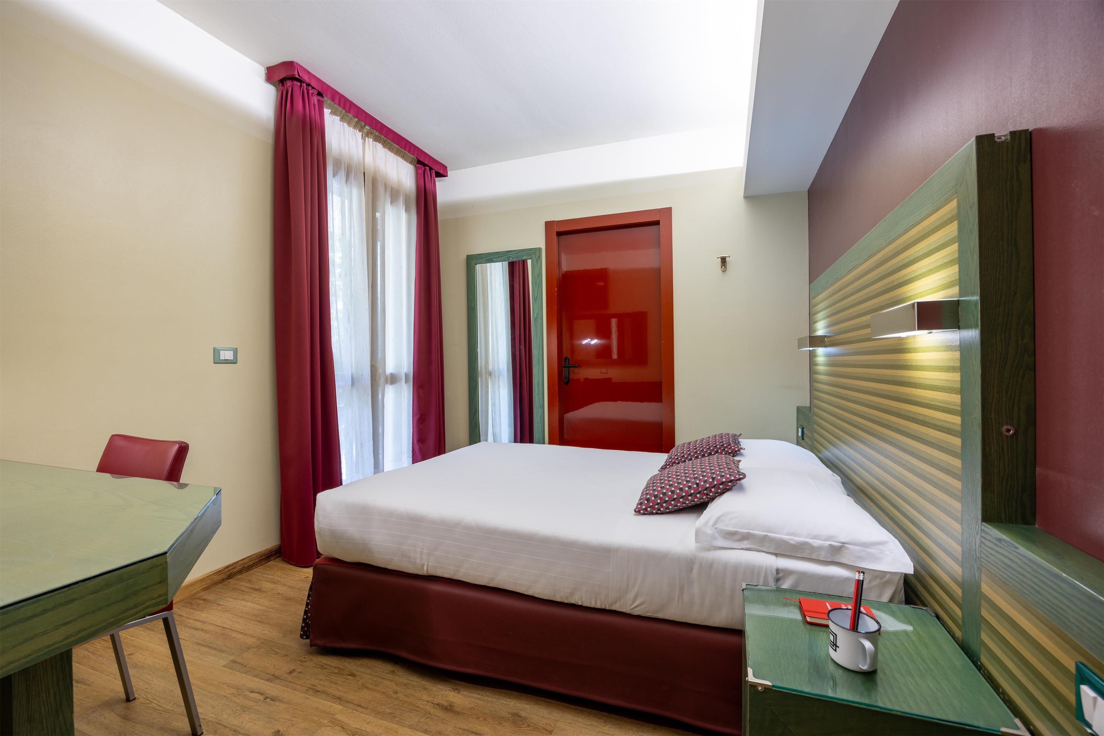 A hotel room with a bed, a desk, a chair, a nightstand, and a window with curtains.