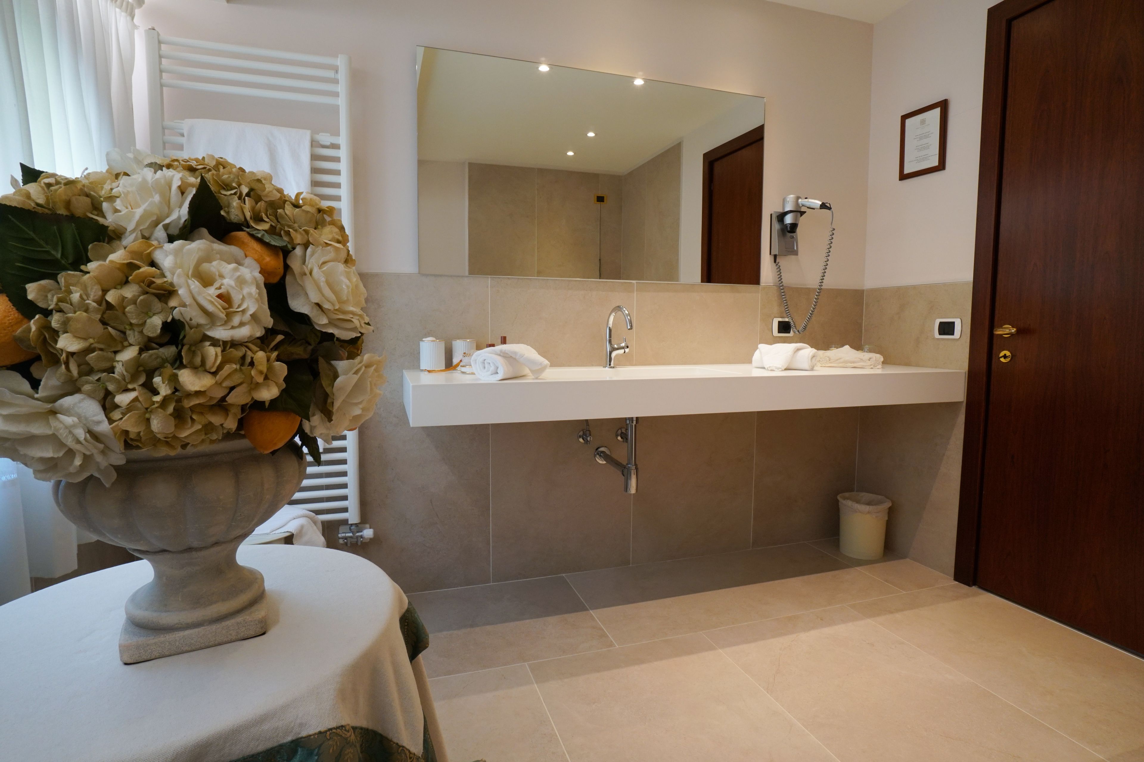 Une salle de bain moderne avec un grand miroir, un lavabo et une douche. On aperçoit un vase rempli de fleurs sur une table au premier plan.
