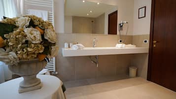 Une salle de bain moderne avec un grand miroir, un lavabo et une douche. On aperçoit un vase rempli de fleurs sur une table au premier plan.