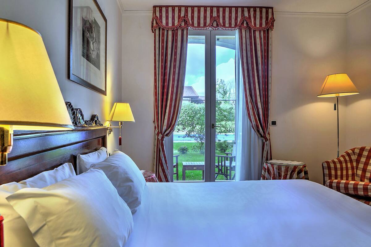 A hotel room with a large bed, a wooden headboard, a small table, a chair, and a view of the outdoors through a glass door.