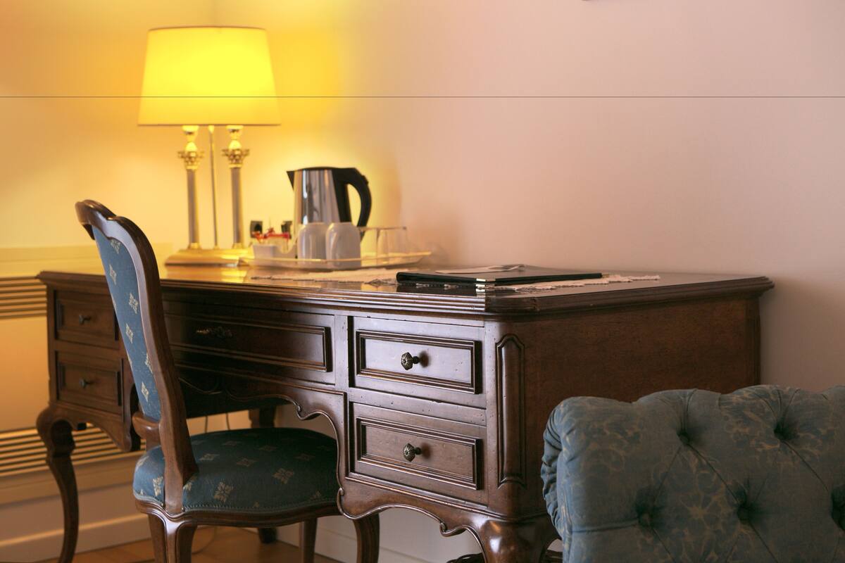A wooden desk with a lamp, a coffee pot, and a chair with a cushion.