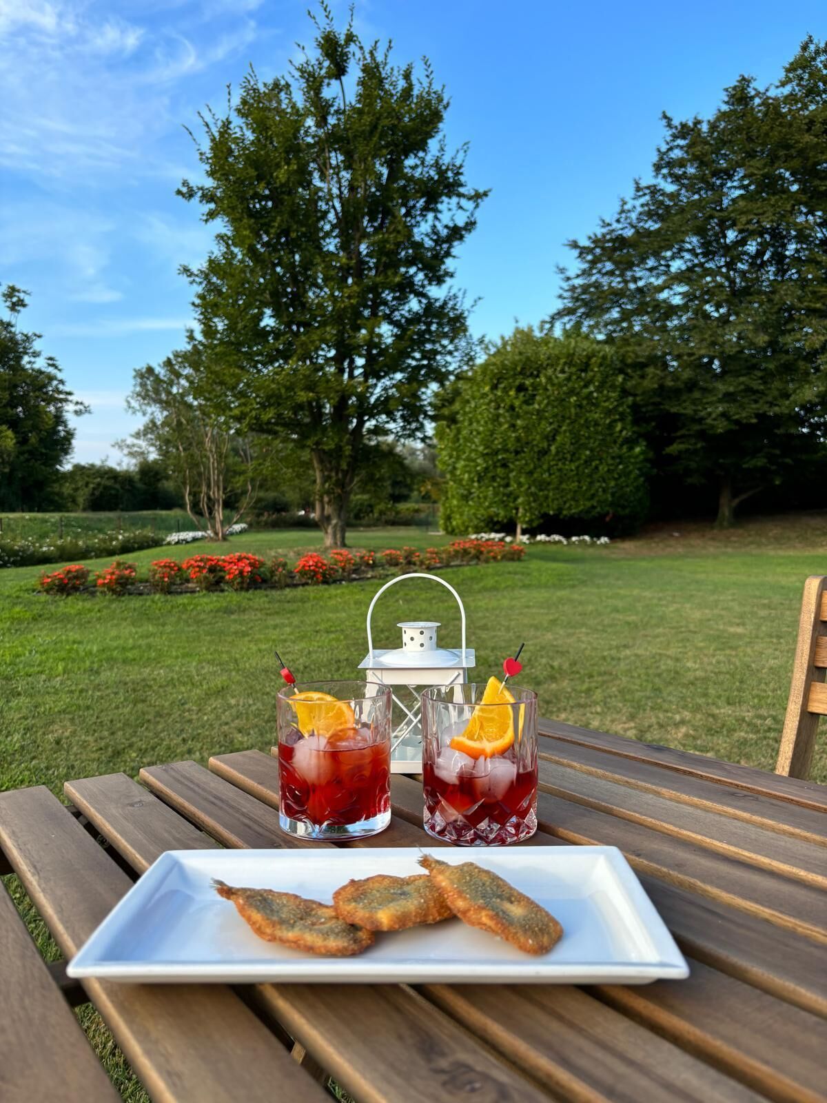 A picnic setup with two drinks, a plate of fried fish, and a lantern on a wooden table in a park.