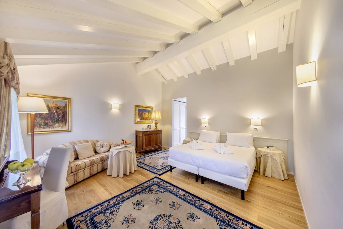 A bedroom with a bed, two bedside tables, a nightstand, a sofa, a small table with fruit, and a painting on the wall.