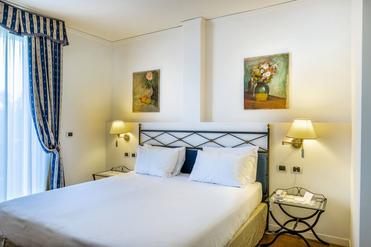 A neatly made bed with white linens, two wall-mounted lamps, and two paintings depicting flowers.