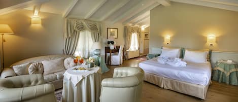 Habitación amplia con una cama grande, un sofá, una silla y una mesita. Hay dos lámparas de pared y una ventana con cortinas.