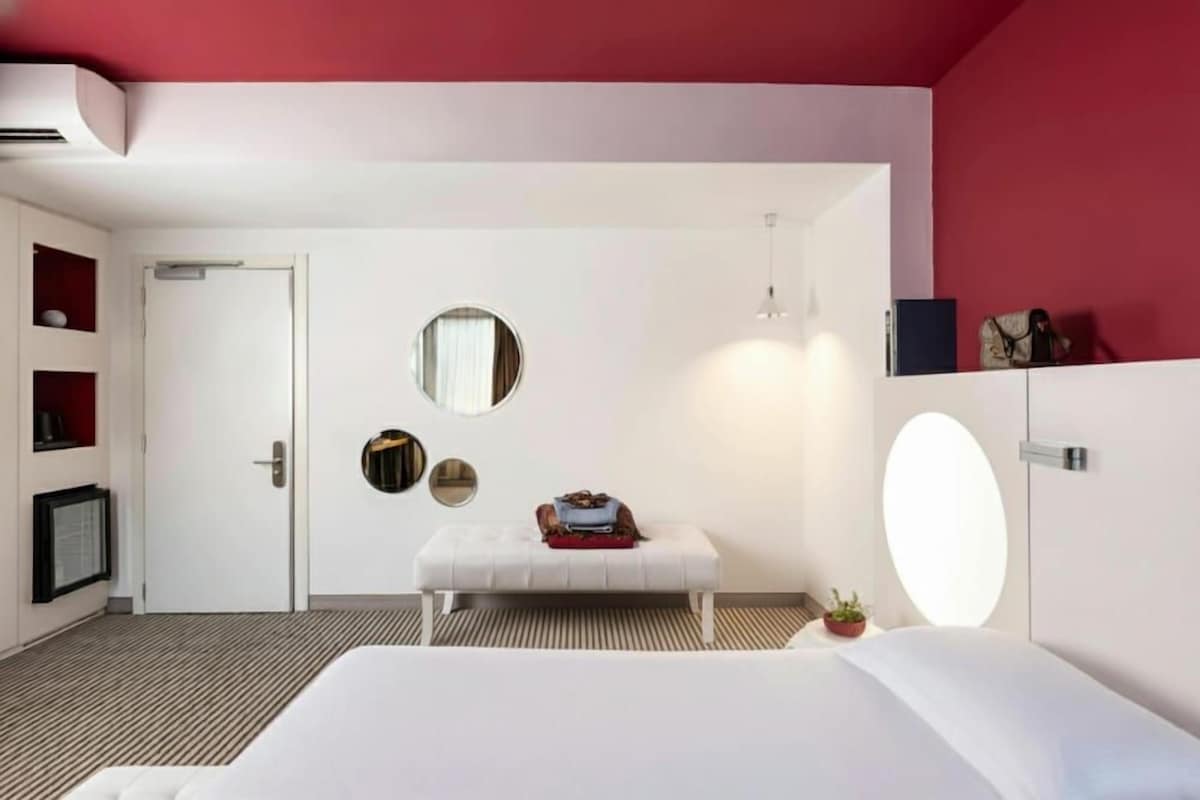 A modern hotel room with a large bed, a round mirror, a bench with a red bag, and a small potted plant.