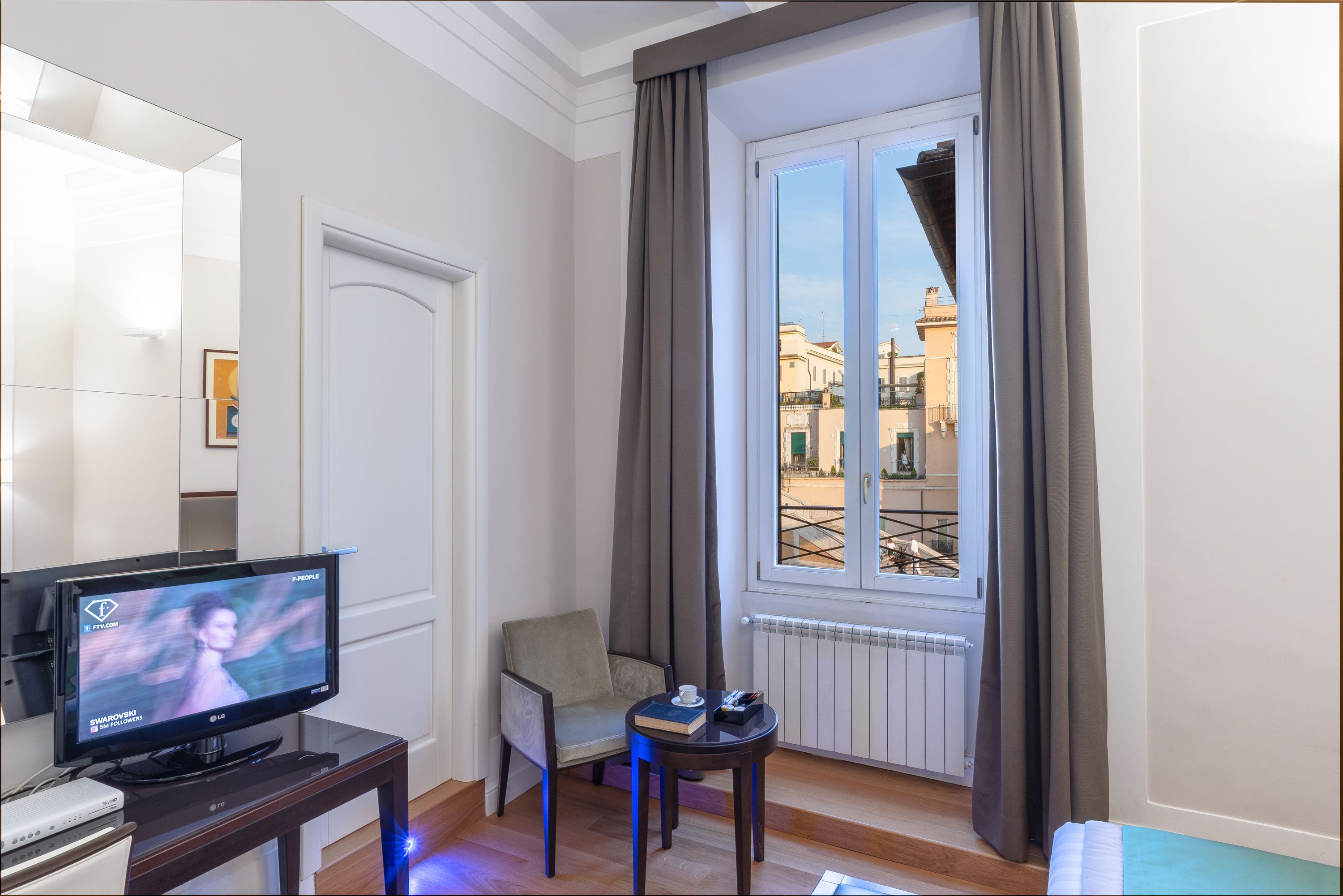 A hotel room with a television, a chair, a small table with a book and a cup, and a window with curtains.