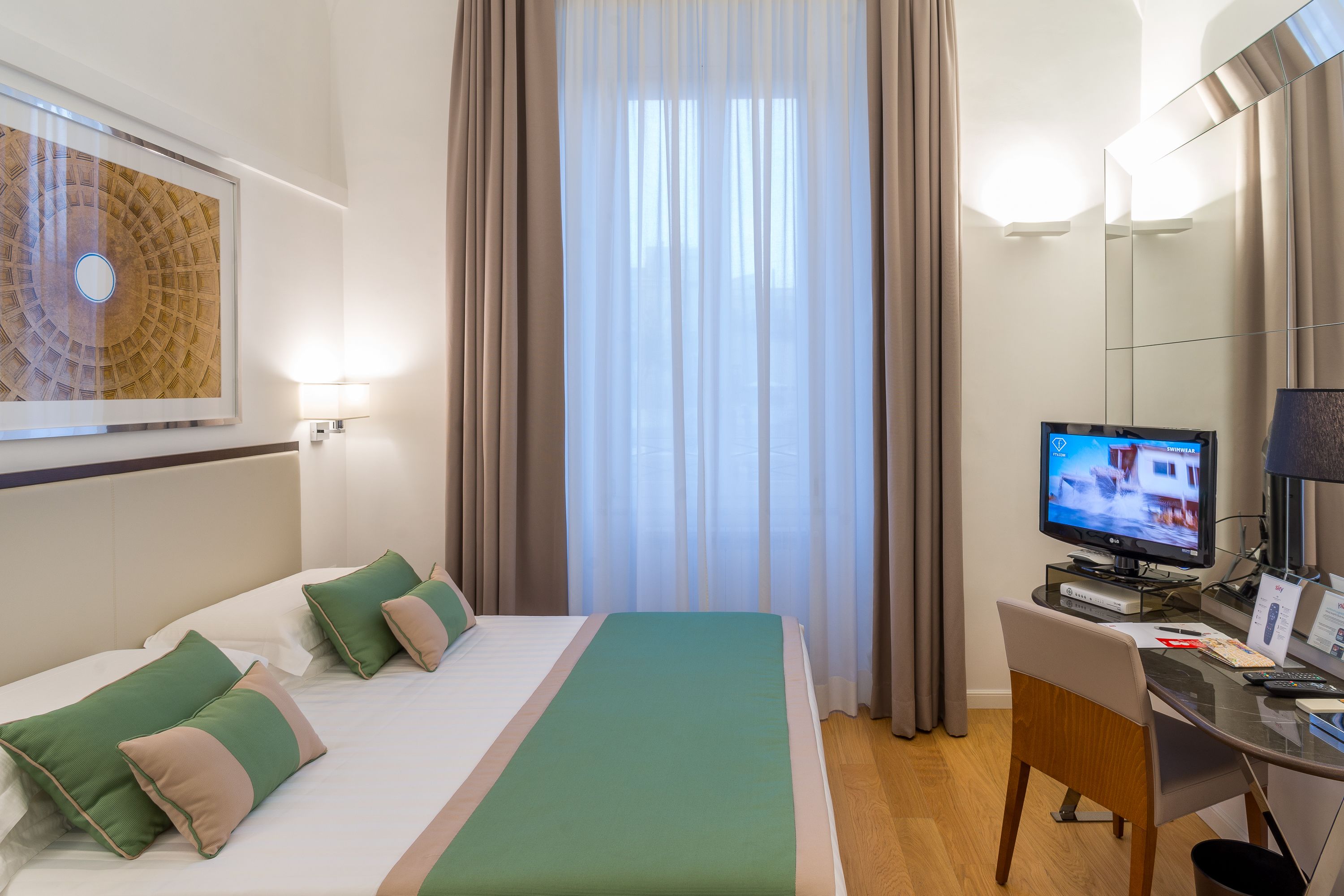 Habitación de hotel con una cama, un escritorio con computadora, un televisor y un espejo grande.