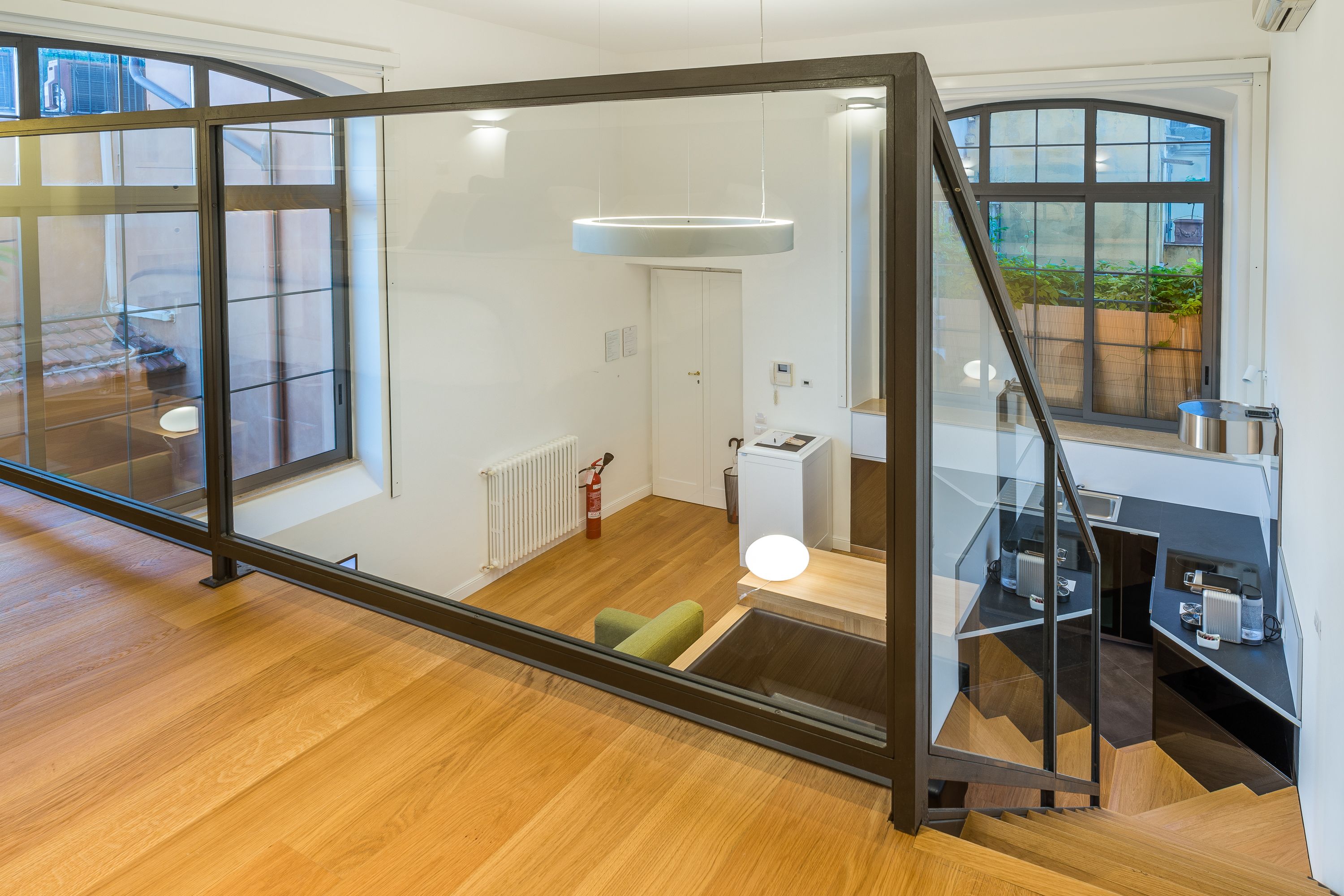 A modern interior with a wooden floor, a staircase with glass railings, and a view of a garden through large windows.