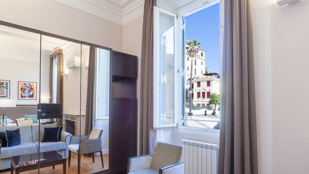 A modern living room with a sofa, armchairs, a coffee table, and a large mirror reflecting the room. A window offers a view of a street with buildings and palm trees. - Internazionale Domus (Rome)