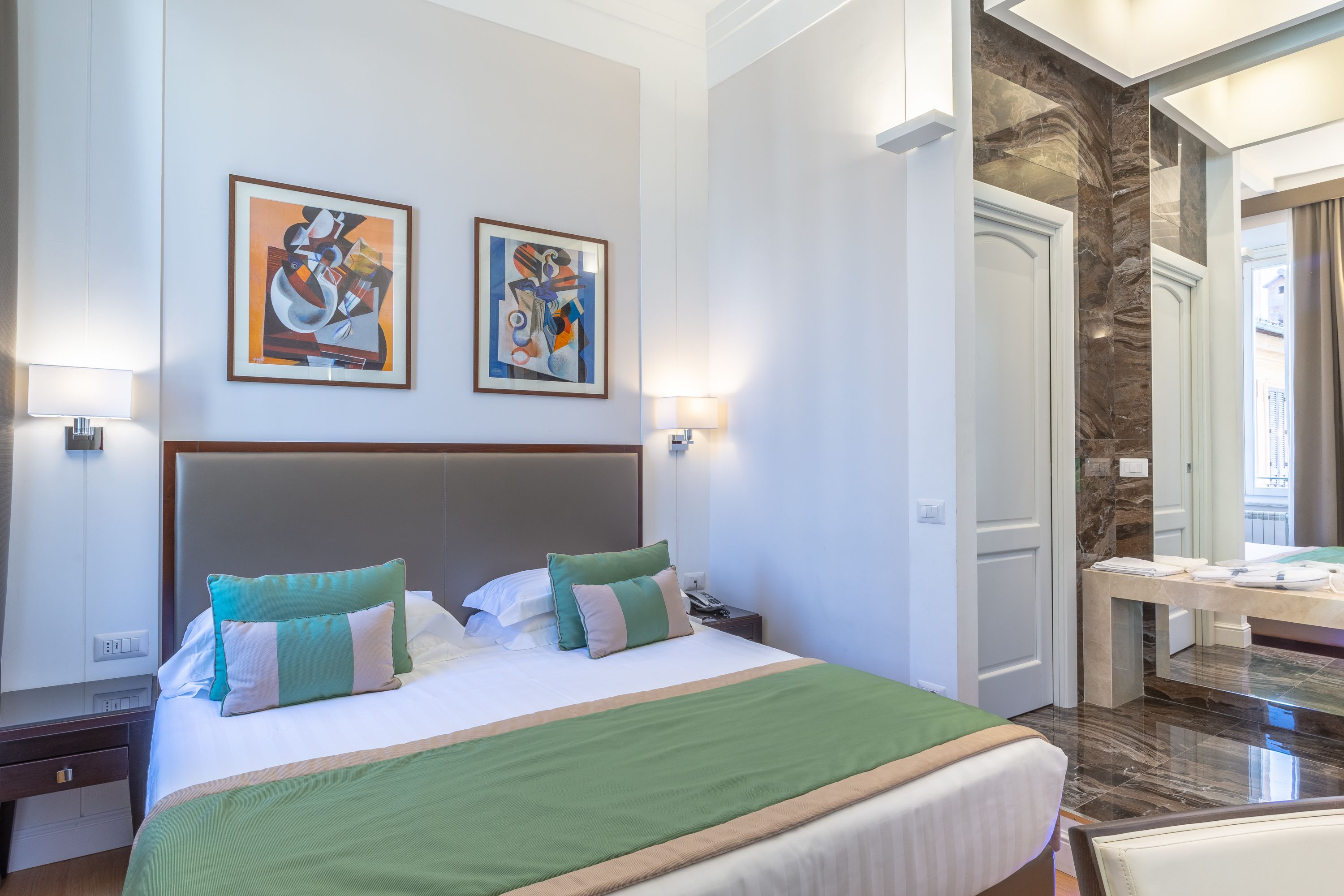 Een hotelkamer met een groot bed, twee aan de muur bevestigde schilderijen, een nachtkastje en een badkamer die door een geopende deur zichtbaar is.