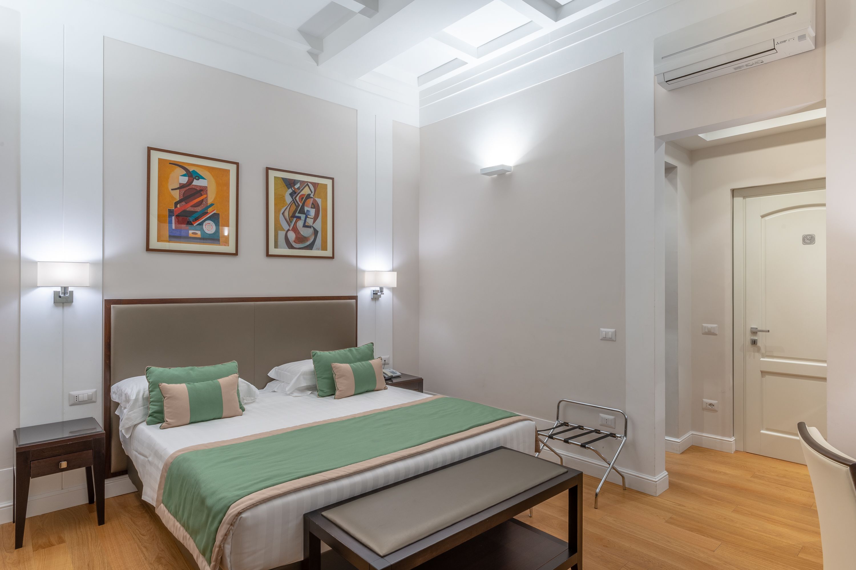 Habitación de hotel ordenada con una cama grande, mesitas de noche, un banco y dos obras de arte colgadas en la pared.