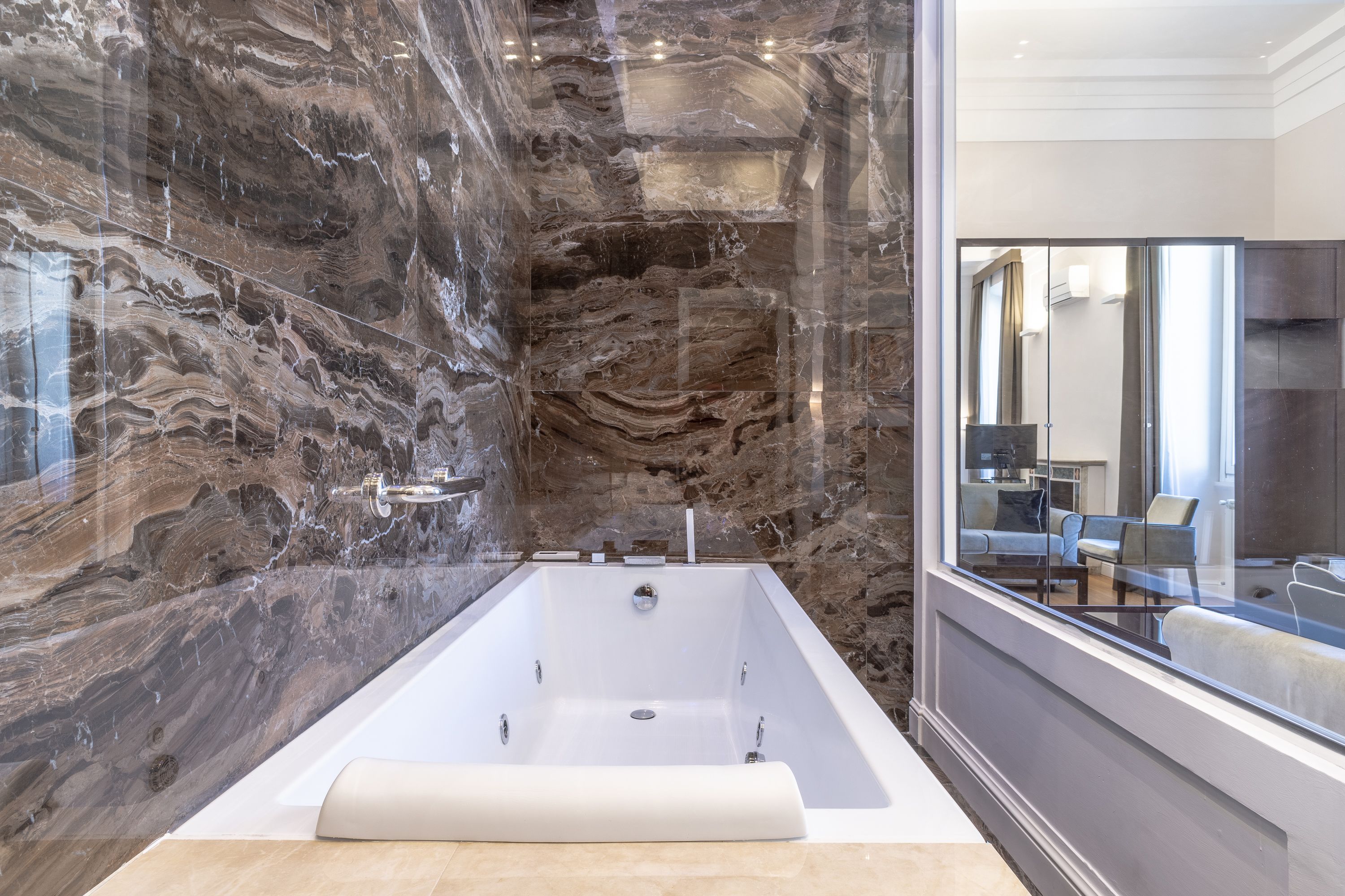 A modern bathroom with a marble wall, a large bathtub, and a view of a living area through a glass partition.