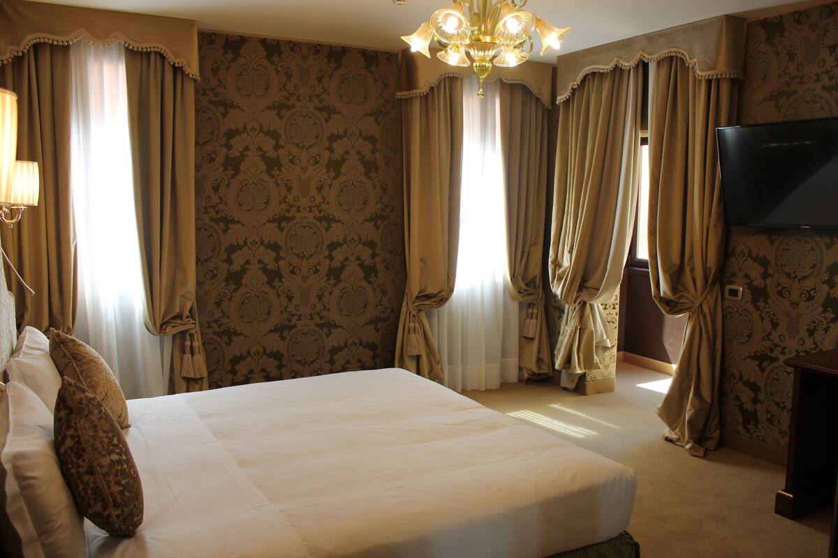 Habitación de hotel con una cama grande, cortinas, una araña y un televisor en la pared.