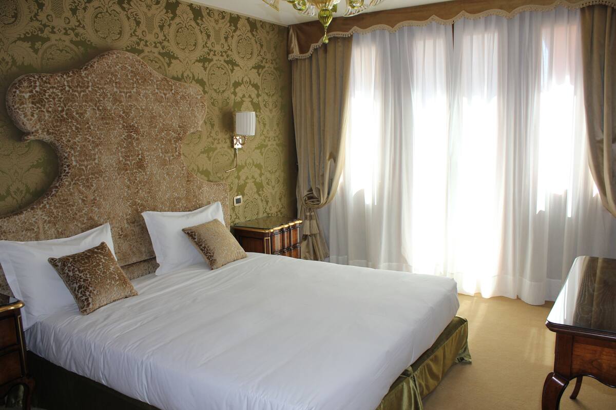Una cama grande con sábanas blancas y dos almohadas decorativas, una mesita de noche con lámpara y una mesa de madera.