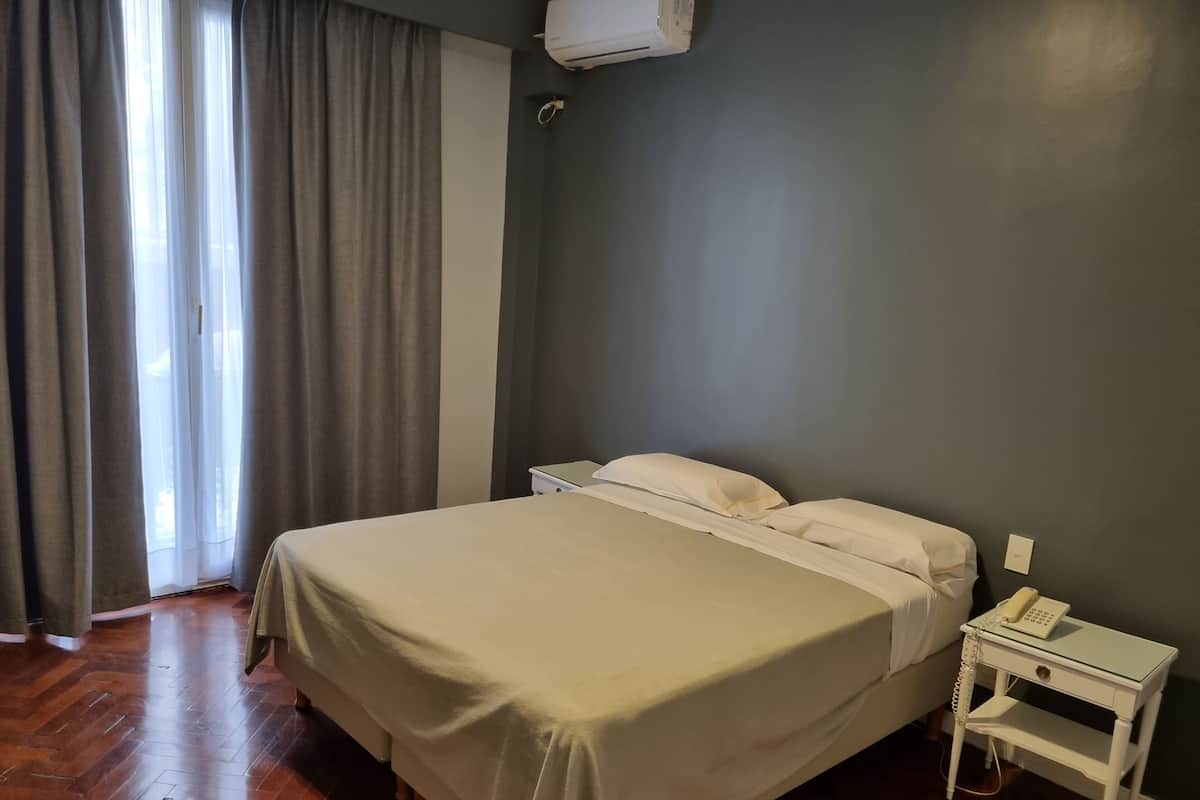A hotel room with a bed, a bedside table with a phone, and a window with curtains.