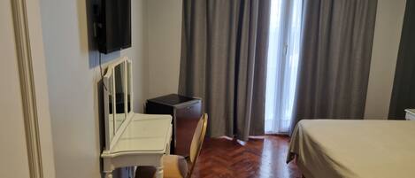 A hotel room with a bed, a desk with a chair, a television mounted on the wall, and a window with curtains.