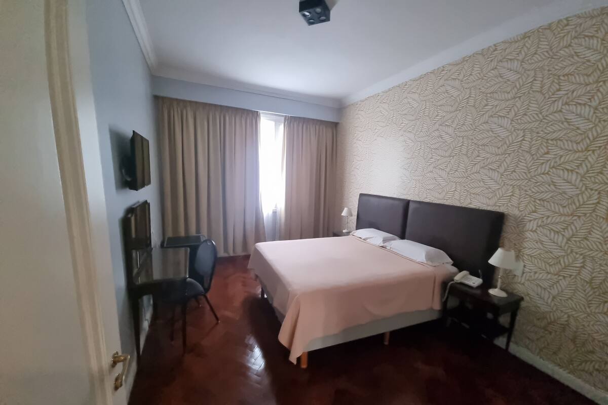 A hotel room with a bed, a desk, a chair, a nightstand, and a lamp.
