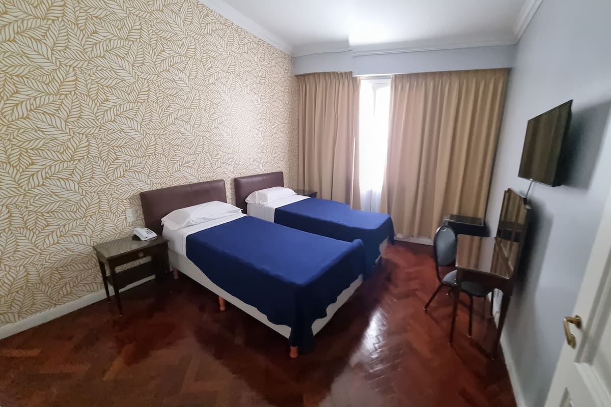 A hotel room with two beds, a desk, a chair, a TV, and a window with curtains.