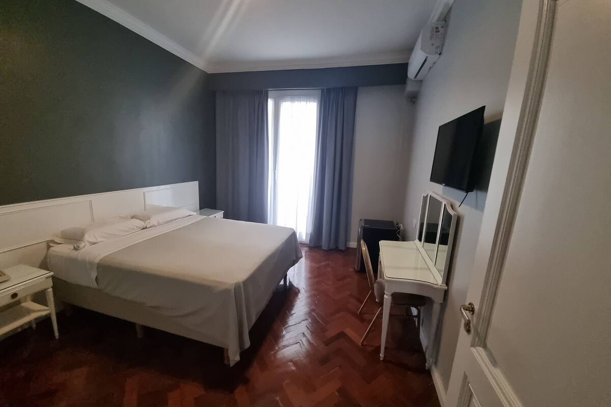 A hotel room with a bed, bedside table, a desk with a chair, a television, and a window with curtains.
