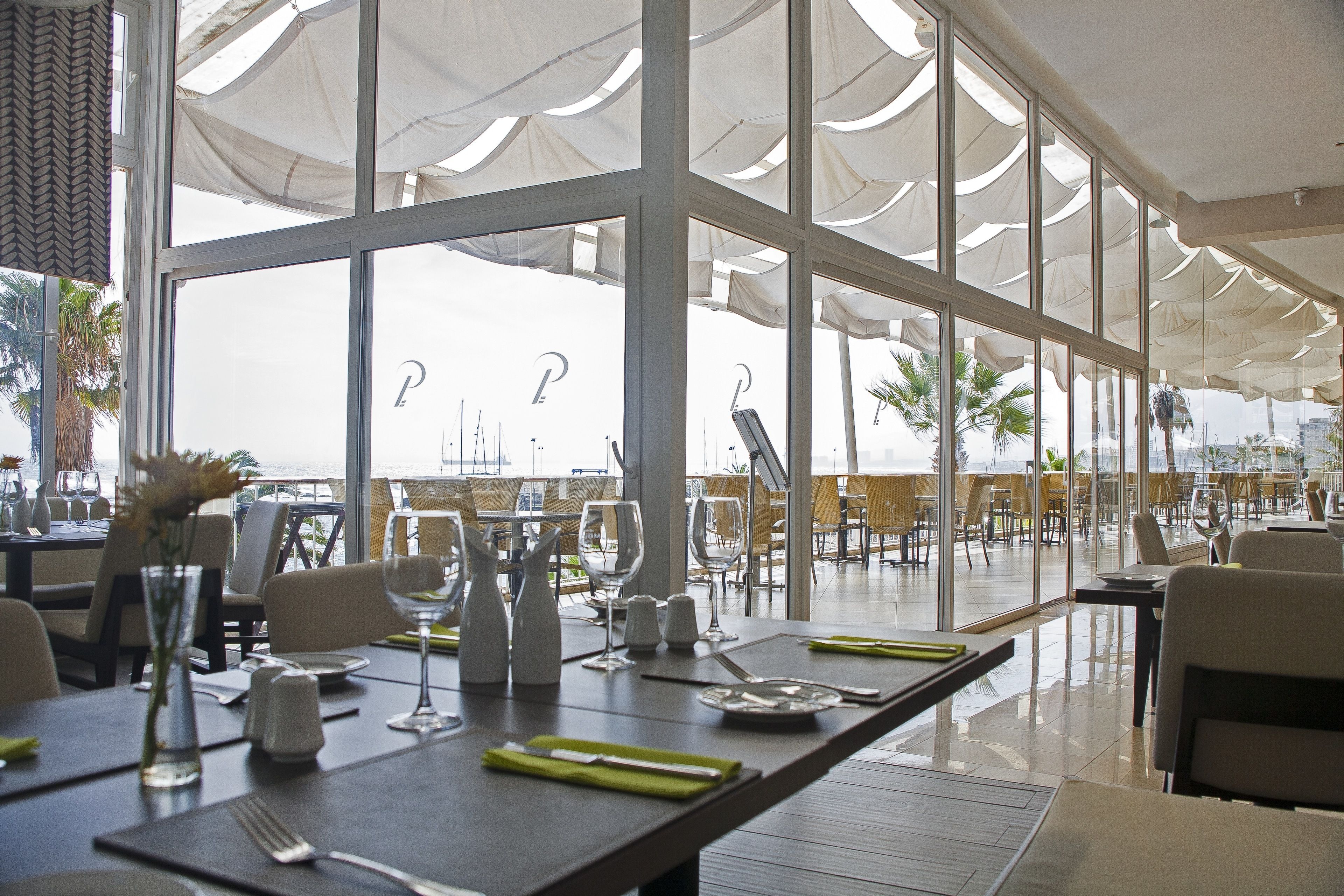 Um restaurante com vista para o oceano, grandes janelas e mesas bem organizadas com talheres e guardanapos.