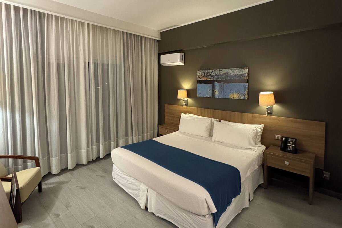 A hotel room with a large bed, two bedside lamps, a wooden headboard, a painting above the bed, a telephone on the nightstand, and a chair by the window.