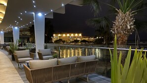 Un área de estar al aire libre con sofás y sillones de mimbre, una barandilla de cristal con vistas a un cuerpo de agua y una vista panorámica de la ciudad de noche.