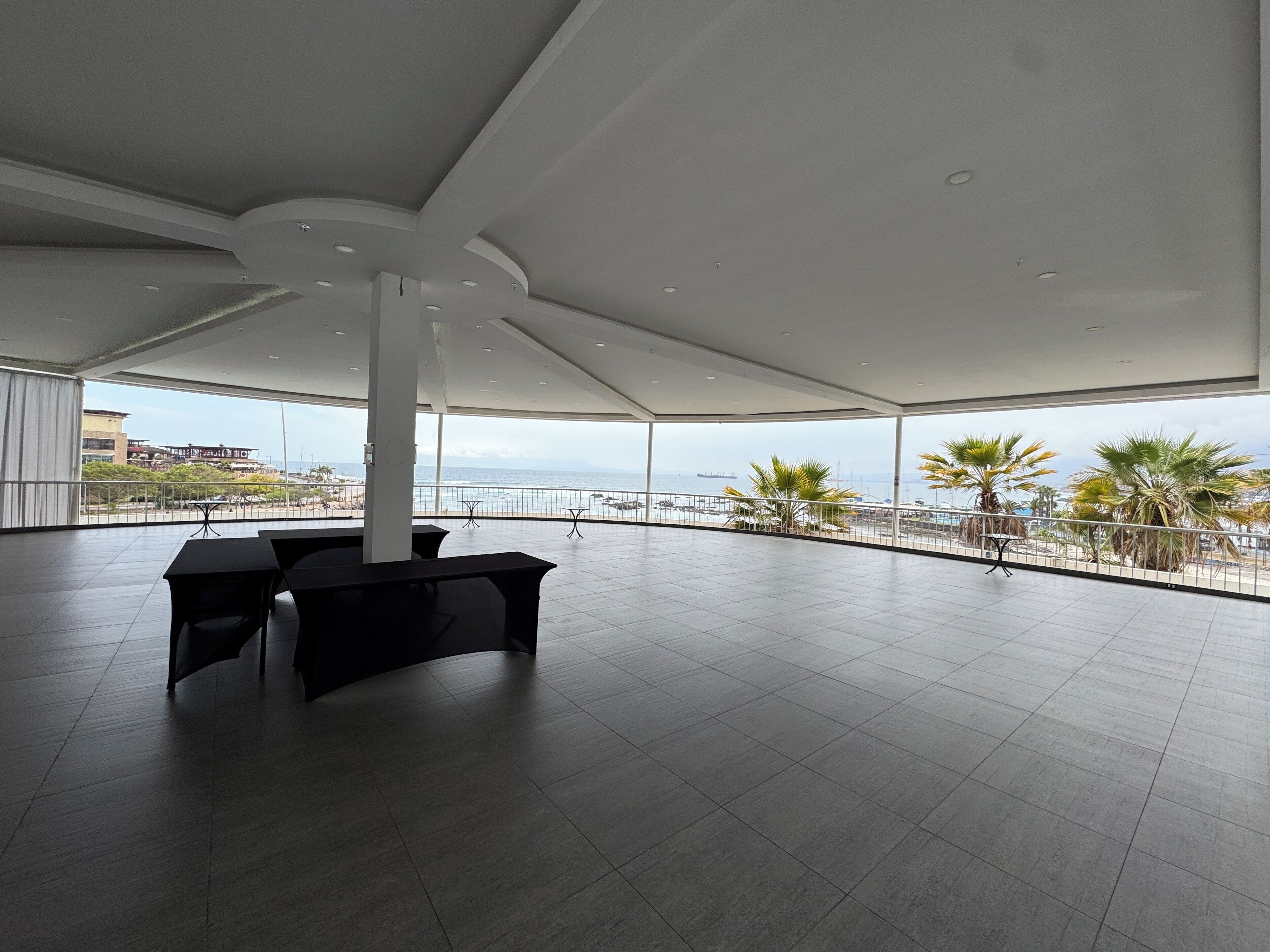 Un espacio interior amplio con techo alto, grandes ventanales que ofrecen vistas al mar y palmeras, y algunas mesas y sillas.