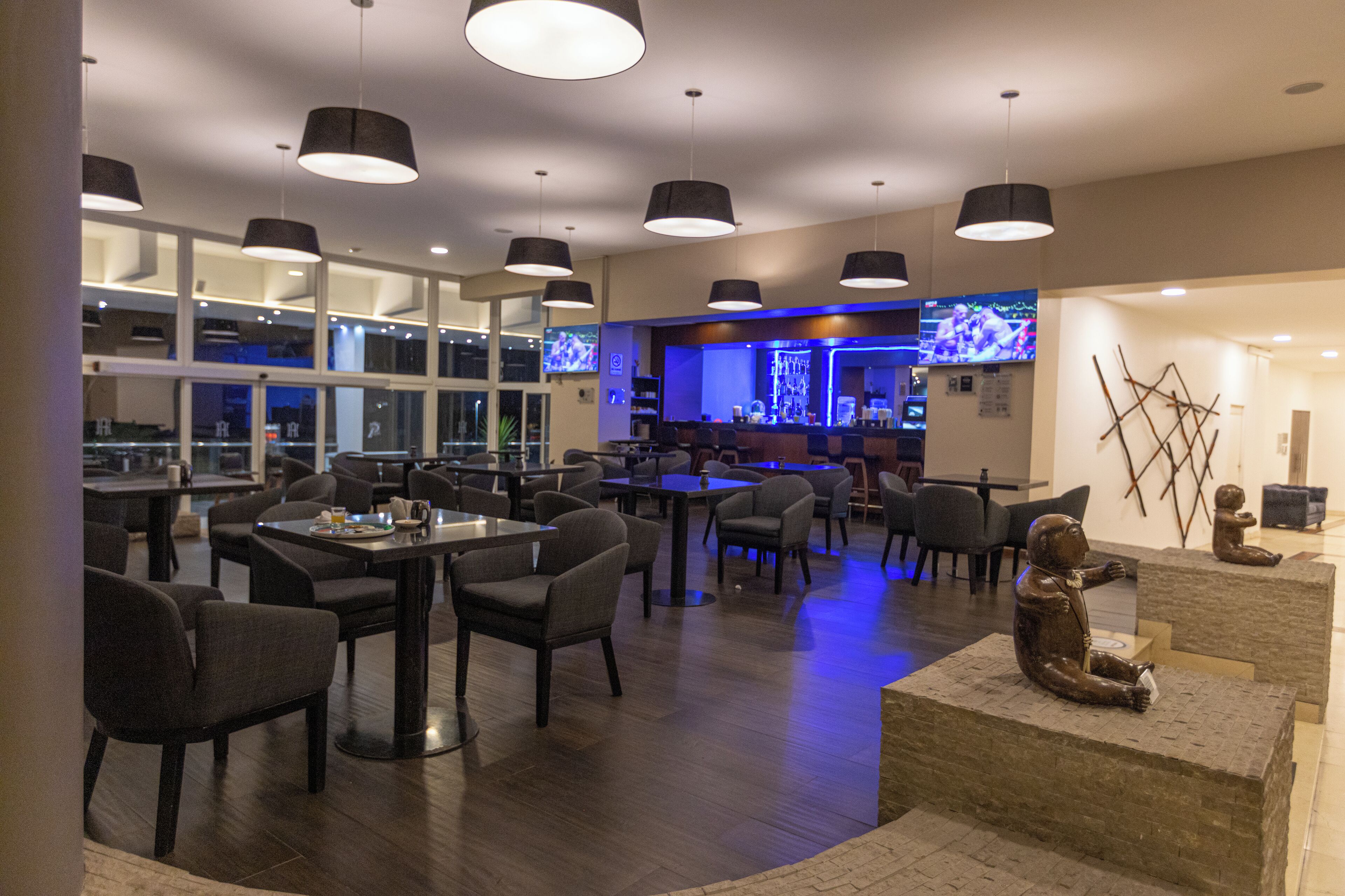 Un restaurante moderno con varias mesas y sillas, una zona de bar con una pantalla de televisión y una escultura decorativa sobre un pedestal.