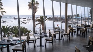 Un restaurante con mesas y sillas dispuestas para comer, amplias ventanas que ofrecen vistas a la playa y al océano, y una zona de piscina visible a través de las ventanas.
