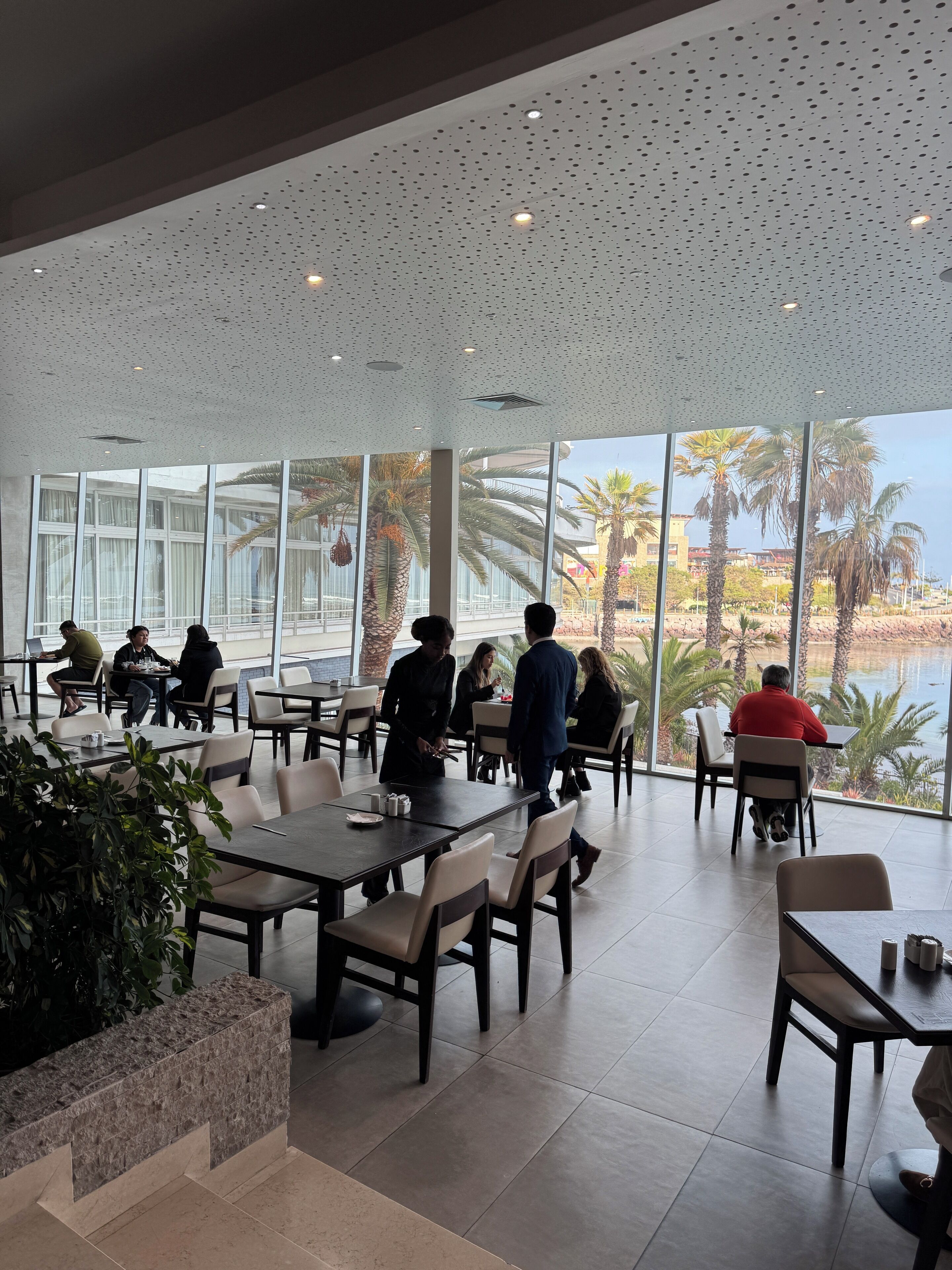 Een ruime eethoek binnen, met grote ramen en uitzicht op palmbomen en een waterpartij. Tafels en stoelen staan klaar voor diners.