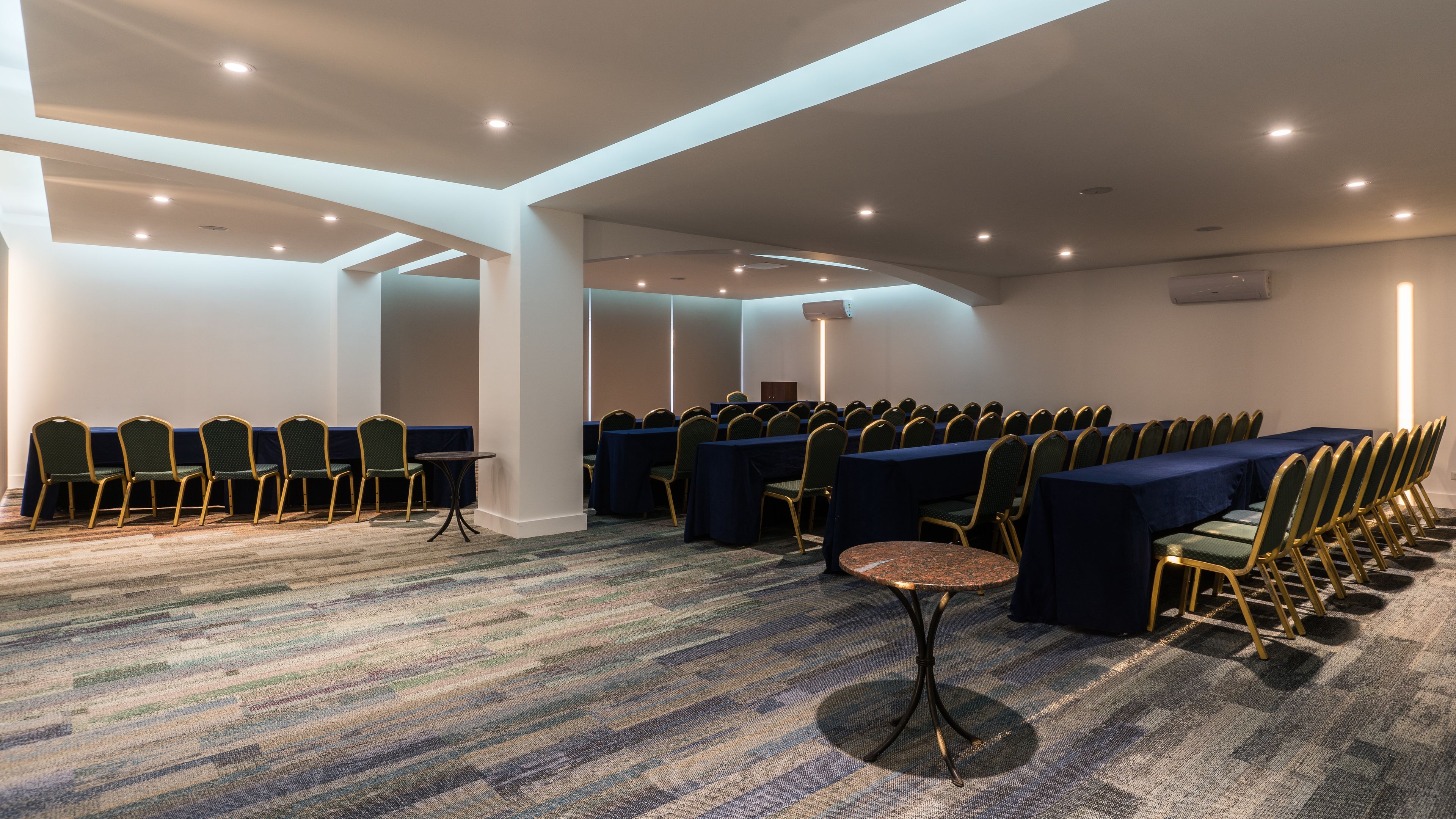 A conference room with rows of tables and chairs, carpeted flooring, and recessed ceiling lighting.
