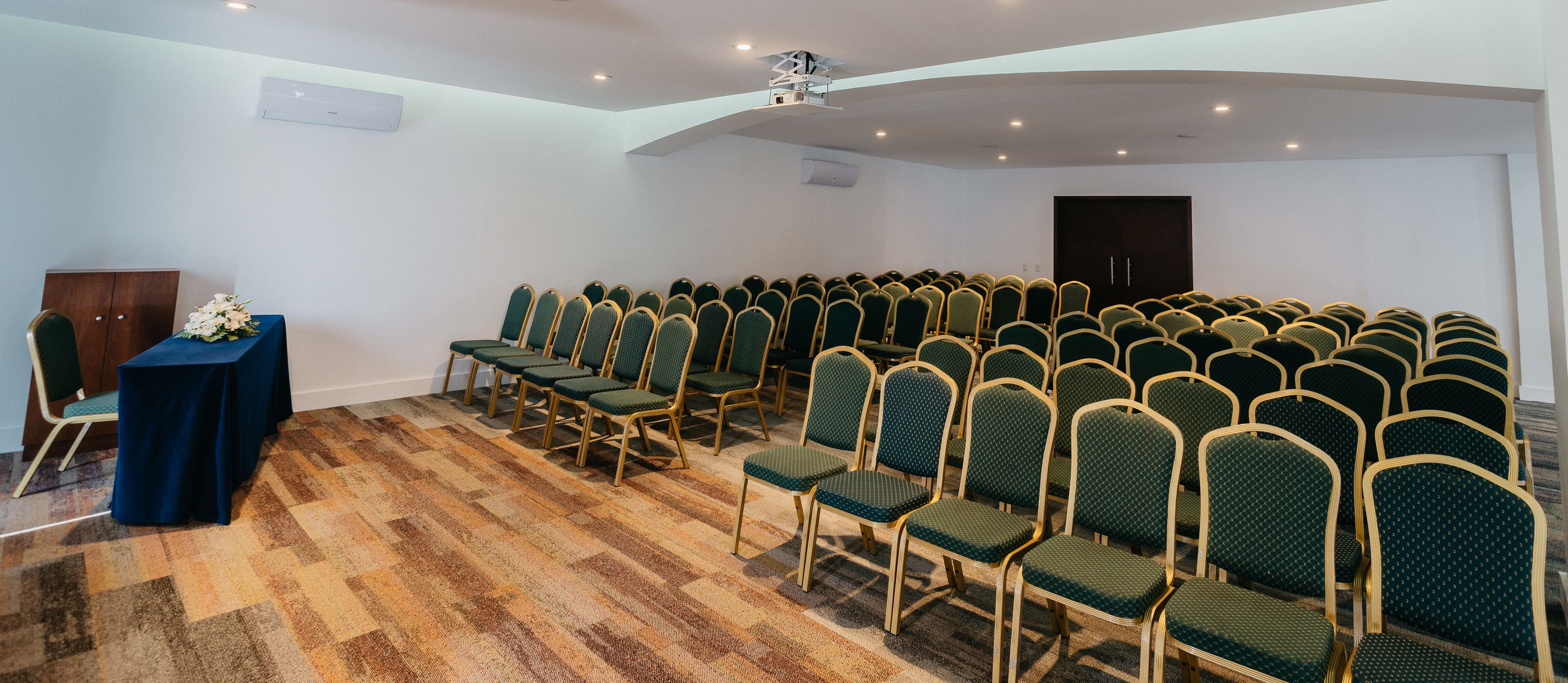 A conference room with rows of green chairs, a blue table with a floral arrangement, and a wooden cabinet.